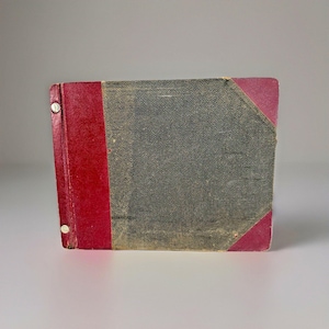 May include: A vintage book with a red spine and corners, and a dark gray cover. The book is bound with metal rivets. The cover shows signs of wear and age, with a textured appearance.