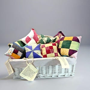 May include: A white wicker basket filled with colorful, patchwork pillows. The pillows feature various patterns and colors, including red, green, blue, and yellow. Each pillow is unique, with a mix of fabrics and designs. The basket has a checkered lining and tags.