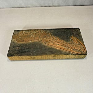 May include: A rectangular wooden printing block with a copper design. The design features a detailed illustration of a building and the cursive text "Santa Barbara, California". The wood is a light brown color, and the copper design is a darker brown.