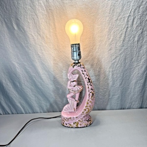 May include: A ceramic table lamp featuring a pink, curved, abstract base with a sculpted figure of a dancer in the center. The base is decorated with gold-colored flecks and supports a single exposed light bulb at the top with a black power cord.
