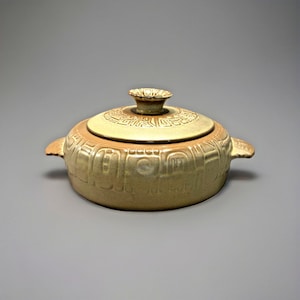 May include: A tan ceramic casserole dish with a lid. The dish has a round shape with two handles and a lid with a small knob. The dish and lid are decorated with embossed geometric patterns.