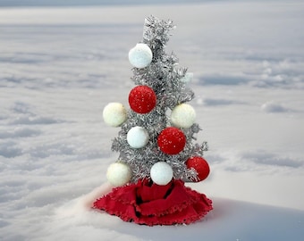 May include: A small silver artificial Christmas tree decorated with red and white ornaments, sitting on a red ruffled fabric base in a snowy landscape.