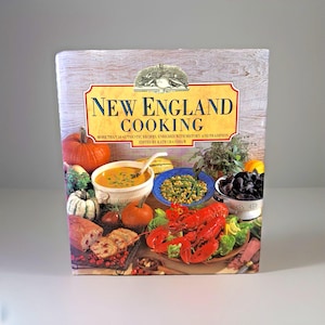 May include: A cookbook titled "New England Cooking" with a cover featuring a variety of dishes, including a lobster, soup, and vegetables. The book's title is in large, gold lettering, with the subtitle "More than 50 Authentic Recipes, Enriched with History and Tradition."