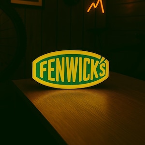 以下が含まれることがあります： 緑色の文字で「FENWICK'S」と書かれた黄色の楕円形の光る看板。木製の表面に置かれ、背景には山型のネオンライトと額縁に入った写真があります。 全体的に暗く、暖色系の色合いです。