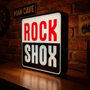 Può includere: Un'insegna quadrata e illuminata con le parole "ROCK SHOX" in grassetto, rosse e bianche. L'insegna ha una cornice nera e poggia su una superficie di legno. Un'insegna "MAN CAVE" è visibile sullo sfondo, suggerendo un tema di cultura automobilistica o motociclistica.