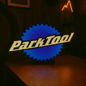 Może przedstawiać: Podświetlany szyld z napisem "Park Tool" w stylizowanej czcionce. Znak ma niebieskie, zębate tło i stoi na drewnianej powierzchni. W tle neon w kształcie góry i oprawiony obraz.