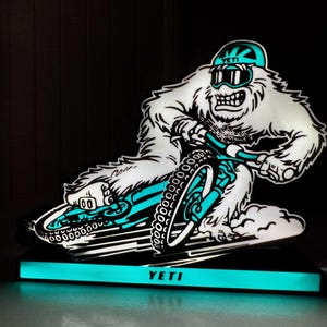 May include: A glowing Yeti mountain bike sign. The white Yeti figure is riding a teal and black mountain bike. The sign has a teal base with the word "YETI" in white. The background is black.
