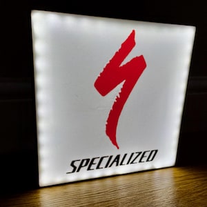 May include: A white, illuminated sign with the word "SPECIALIZED" in black text. A red, stylized lightning bolt logo is centered above the text. The sign is lit from within, creating a bright, glowing effect.