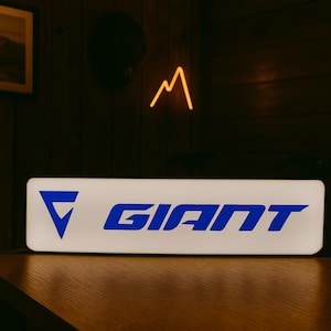 May include: A rectangular, illuminated sign with the word "GIANT" in blue capital letters. The sign has a white background and a blue triangle logo. The sign is on a wooden surface, with a dark background.