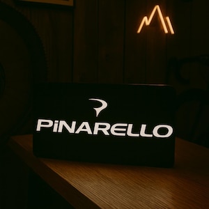 May include: A black rectangular object with the white text "PINARELLO" and a stylized logo. The object is on a wooden surface, with a neon mountain-shaped light in the background.