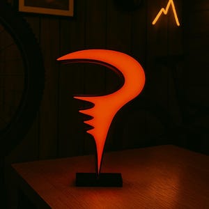 May include: An illuminated orange sculpture with a unique, stylized shape, set on a black base. The sculpture is on a wooden table, with a mountain-shaped neon light in the background. The overall theme is modern art.
