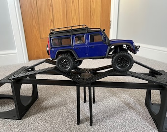 May include: A blue toy Land Rover Defender driving over a black plastic ramp obstacle course. The ramp is made of interlocking pieces and has a bridge section.