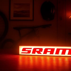 以下が含まれることがあります： 太字の赤い文字で「SRAM」という単語が表示された、長方形の照明付きサイン。サインは内側から照らされ、暖かいオレンジ色の光を放っています。背景には自転車の車輪、ヘルメット、ネオンの山の輪郭が含まれています。