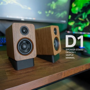 May include: Two wooden speaker stands with black bases. The stands are on a wooden desk. The text "D1 SPEAKER STAND DESIGN-1 SIMPLE MINIMAL" is visible in the image.