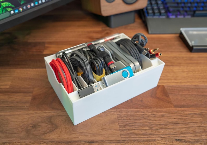 May include: A white plastic organiser with multiple compartments filled with various cords, cables, and a blue USB drive. The organiser is sitting on a wooden surface.