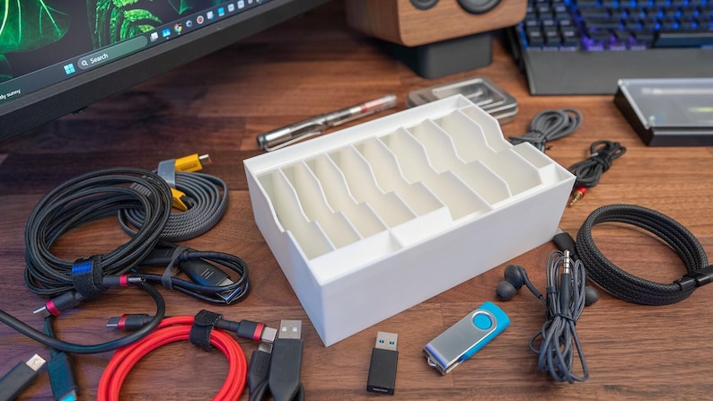 May include: A white plastic cable organiser with multiple compartments, surrounded by various cables, including black, grey, red, and yellow cables, a pair of blue earbuds, and two USB drives.