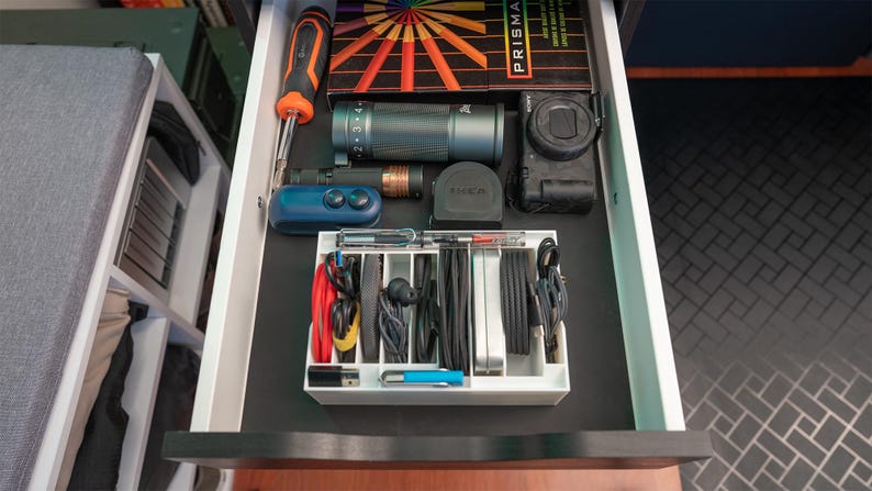 May include: An open drawer with a white organiser containing various cords, cables, and a blue pen. A camera, a flashlight, and a screwdriver are also visible in the drawer.
