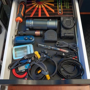 May include: An open drawer containing a variety of electronics and accessories, including a camera, a lens, a remote control, a small device with a screen, and several cables and wires. The drawer is lined with black material.