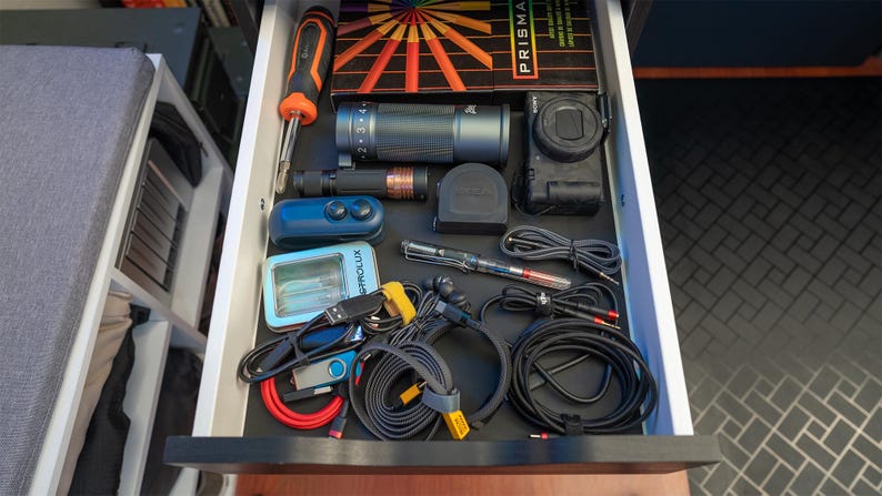 May include: An open drawer containing a variety of electronics and accessories, including a camera, a lens, a remote control, a small device with a screen, and several cables and wires. The drawer is lined with black material.