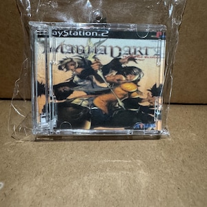 May include: A miniature clear plastic case containing a small replica of the MagnaCarta: Tears of Blood video game cover art for the PlayStation 2, featuring illustrated characters in a battle scene, enclosed in a sealed transparent plastic bag.