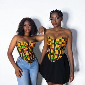 May include: Two strapless corset tops featuring a vibrant geometric Kente-inspired pattern in yellow, green, red, and black. Each corset has a central black zipper closure and vertical black boning details. One is paired with blue jeans, the other with a black skirt.