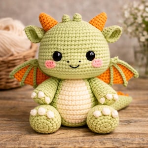 May include: A handmade crochet dragon toy in shades of green, orange, and cream. The dragon has large black eyes, pink cheeks, and orange horns and wings. The dragon is sitting on a wooden surface.