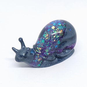 May include: A decorative snail ornament with a dark grey body and a shell filled with colourful glitter. The glitter includes shades of blue, green, purple, and gold. The snail has two small antennae and is set against a white background.