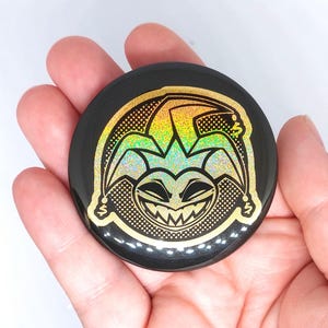 May include: A circular, black badge featuring a holographic jester design. The jester's face and hat are outlined in gold, with a rainbow-coloured fill. The badge has a glossy finish and is likely a pin.