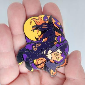 May include: A purple and orange enamel pin featuring a cartoon character with a large, toothy grin. The character is wearing a purple and black hat and is holding a smaller, orange character with a yellow moon behind them.