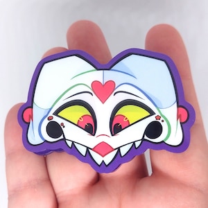 May include: A colorful sticker featuring a cartoon character's face with white hair, yellow eyes, and a red heart. The sticker has a purple border and is held in a hand.