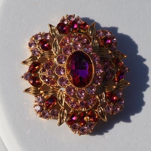 Puede incluir: Broche dorado con una piedra central ovalada de color púrpura oscuro, rodeada de cristales más pequeños, redondos, rosas y rojos. El broche tiene un diseño floral con detalles de hojas doradas, creando una estética vintage o antigua.