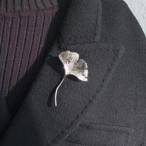 May include: A silver ginkgo leaf brooch pinned to a black fabric background.