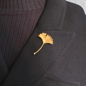 May include: A gold-colored ginkgo leaf brooch pinned to a black jacket.