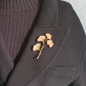 Könnte beinhalten: Eine goldfarbene Brosche in Form eines Ginkgoblatt-Zweigs mit funkelnden Strasssteinen. Die Brosche ist an einem schwarzen Blazer befestigt.