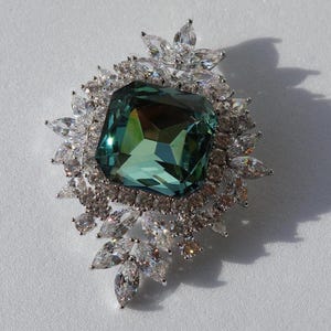 Può includere: Una grande gemma verde smeraldo circondata da un alone di gemme più piccole e trasparenti. Il pezzo è incastonato in argento e presenta un delicato design floreale.