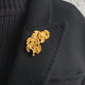 Könnte beinhalten: Eine goldfarbene Brosche in Form eines stilisierten Zweigs mit Blättern. Die Brosche befindet sich auf einem schwarzen Stoffhintergrund.
