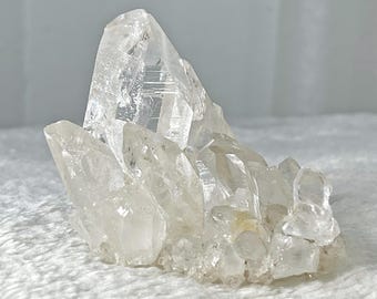 May include: A cluster of clear quartz crystals with a large, pointed crystal in the center. The crystals are arranged in a natural, organic shape.