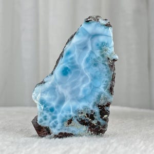 May include: A polished piece of larimar gemstone, showcasing a light blue hue with swirling patterns reminiscent of ocean waves. The stone has a dark brown and black matrix, creating a striking contrast. The overall shape is irregular and natural.