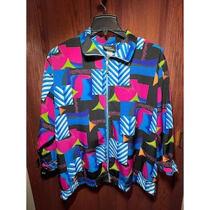 May include: A colorful zip-up jacket with a vibrant geometric pattern. The jacket features a black base with pink, blue, green, and orange shapes, accented by white striped details. It has a stand-up collar and a full-length zipper.