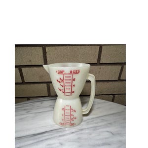 May include: A vintage cream-coloured measuring jug with red markings. The jug is marked with measurements in cups and fluid ounces, with a capacity of 237 ml. It has a handle and a spout. The jug is made of a durable material.
