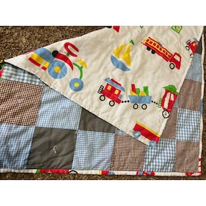 Pode incluir: Um edredom de retalhos com uma parte superior branca com ilustrações coloridas de um triciclo, um veleiro, um carro de bombeiros e um comboio. A parte inferior do edredom é um patchwork de quadrados azuis, castanhos e cinzentos.