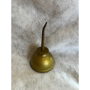 May include: A vintage brass oil can with a curved spout and a rounded base. The can has a tarnished, aged appearance, suggesting it is an antique or a well-used item. The overall design is simple and functional.