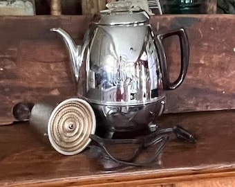 Op de afbeelding: Een glanzende verchroomde elektrische percolator koffiepot met een donkerbruin handvat, een doorzichtige glazen knop op het deksel en een bijpassend intern metalen mandje. Een zwart netsnoer ligt opgerold voor de koffiepot.