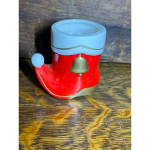 May include: A ceramic boot-shaped vessel featuring a glossy red finish with a white cuff at the top. The boot is decorated with a gold-coloured bell accent on the side and a small white sphere at the toe, finished with a gold-coloured base trim.