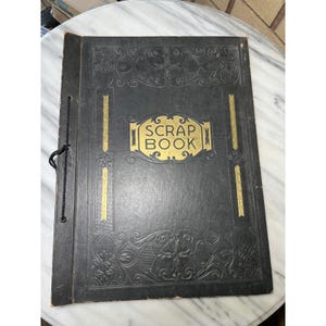 Puede incluir: Un álbum de recortes negro vintage con detalles dorados y las palabras "SCRAP BOOK" en letras doradas. La portada tiene diseños florales en relieve y rayas verticales doradas. Una cuerda negra une las páginas.