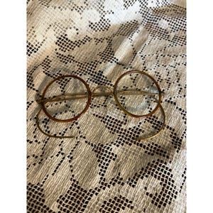 Peut inclure: Lunettes rondes vintage avec une monture en métal doré et des verres teintés marron. Les lunettes ont un design classique, avec un pont et des branches. Les verres sont circulaires et la monture est fine et délicate.
