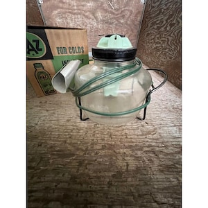 May include: Vintage glass vaporizer with a black lid and a green top. The vaporizer has a metal handle and is wrapped with green tubing. A cardboard box with the text "AZ FOR COLDS" is in the background.