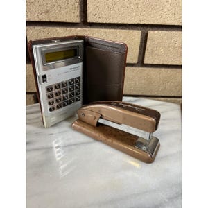 May include: A vintage silver and brown Sharp electronic calculator with a brown leather case and a brown Swingline stapler. The calculator has a digital display and a keypad. The stapler has the text "Swingline 55" on it.