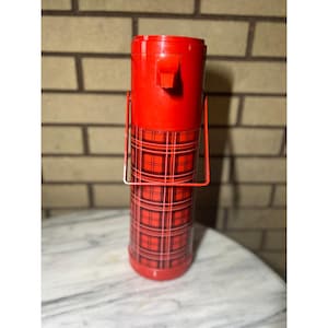 May include: A vintage red thermos with a plaid pattern on the lower half. The thermos has a handle and a spout. The background is a brick wall and a marble-like surface.