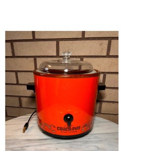 Könnte beinhalten: Ein leuchtend orangefarbener Rival Crock-Pot Slow Cooker mit Glasdeckel. Das Gerät hat schwarze Griffe und einen Bedienknopf. Die Worte "Rival Crock-Pot Slow Cooker" sind auf der Vorderseite zusammen mit dekorativen Bildern aufgedruckt.